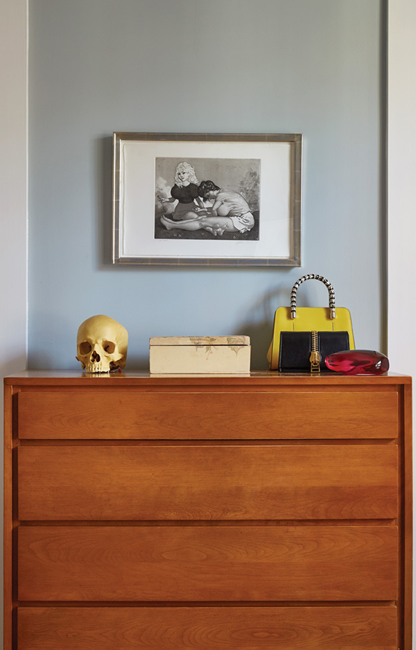 A dresser by Leslie Diamond for Conant Ball, often attributed to Russel Wright, holds several treasures—a music box made from a plastic skull by Douglas Gordon; a glass stone by Arlan Huang; and a vintage handbag. Above it hangs an etching from John Currin’s “Milestones” portfolio. | Photography by MARK ROSKAMS